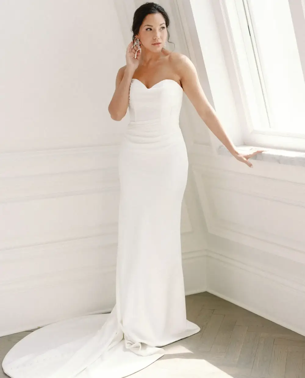 A woman in a strapless white wedding dress stands next to a window, elegantly posing with one hand near her ear.