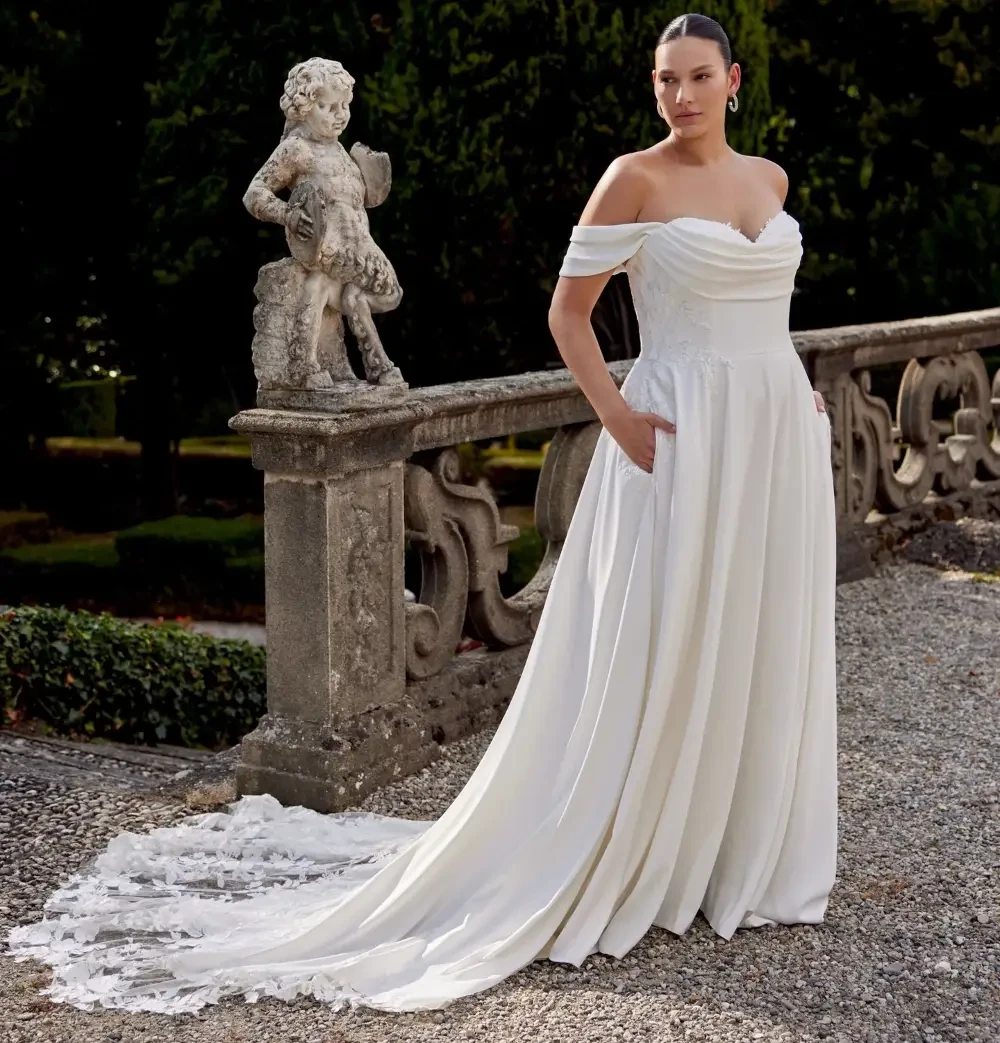 A woman stands elegantly in a white off-the-shoulder wedding dress with pockets, posing in a garden setting beside a statue.