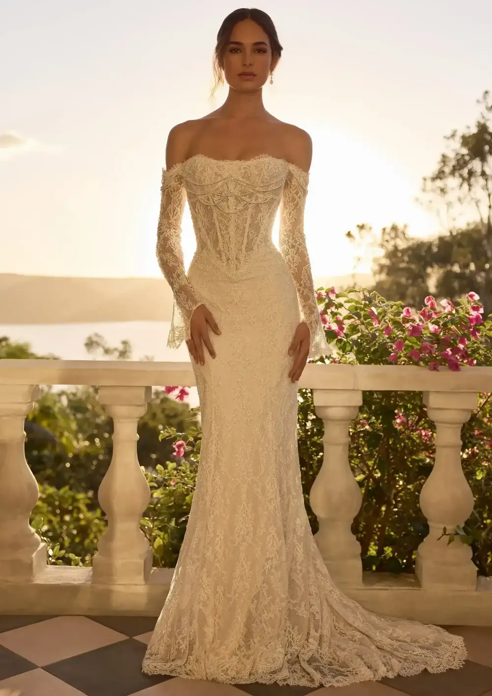 A woman wearing an elegant off-the-shoulder lace gown stands on a balcony, surrounded by flowers and greenery, with a scenic background at sunset.