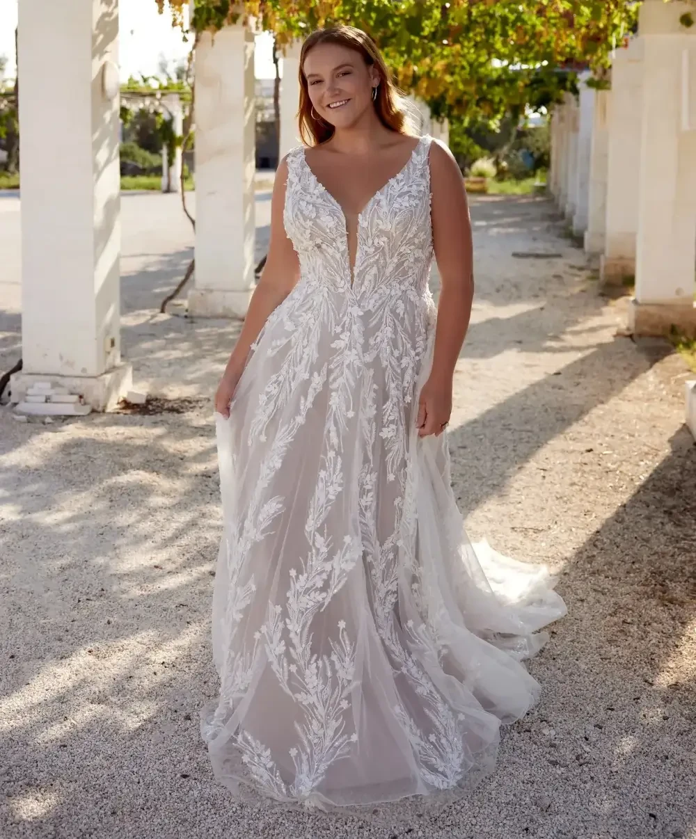 A woman wearing a beautiful wedding dress with intricate floral designs, standing in a sunlit outdoor setting.