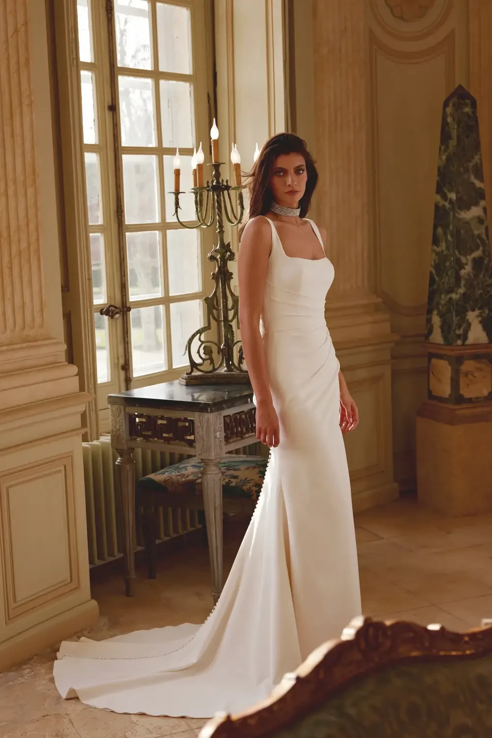 A woman in an elegant white dress stands in a beautifully decorated room with large windows and antique furnishings.