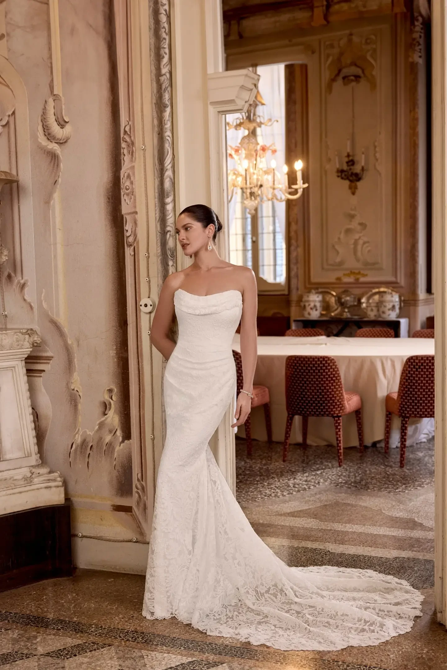 A woman in a strapless, lace wedding gown stands elegantly in a luxurious, ornate room. The mood is sophisticated and serene, with a chandelier in the background.