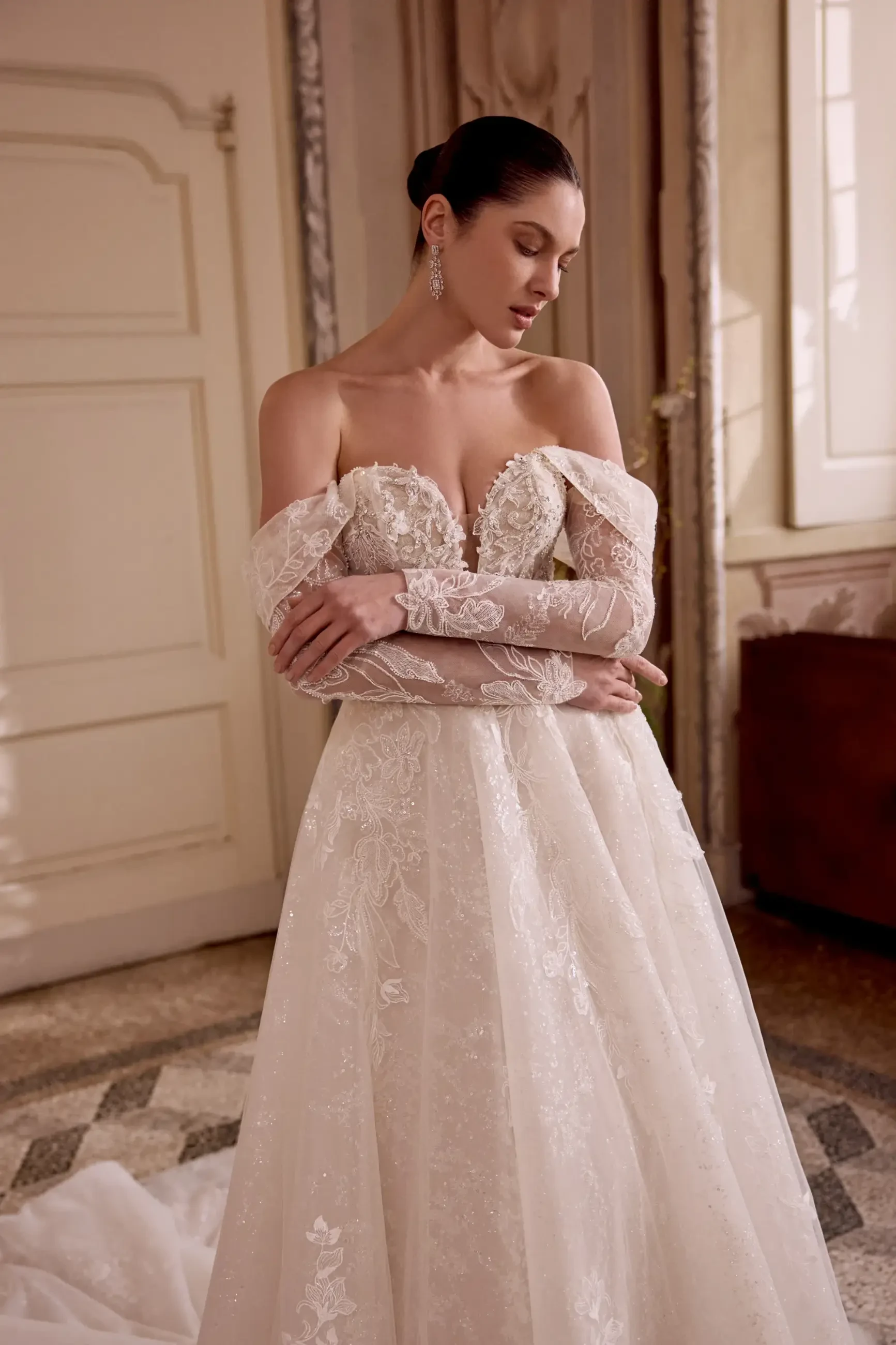 Bride in elegant, off-shoulder lace wedding gown with intricate details. She stands in a softly lit, ornate room, exuding grace and contemplation.