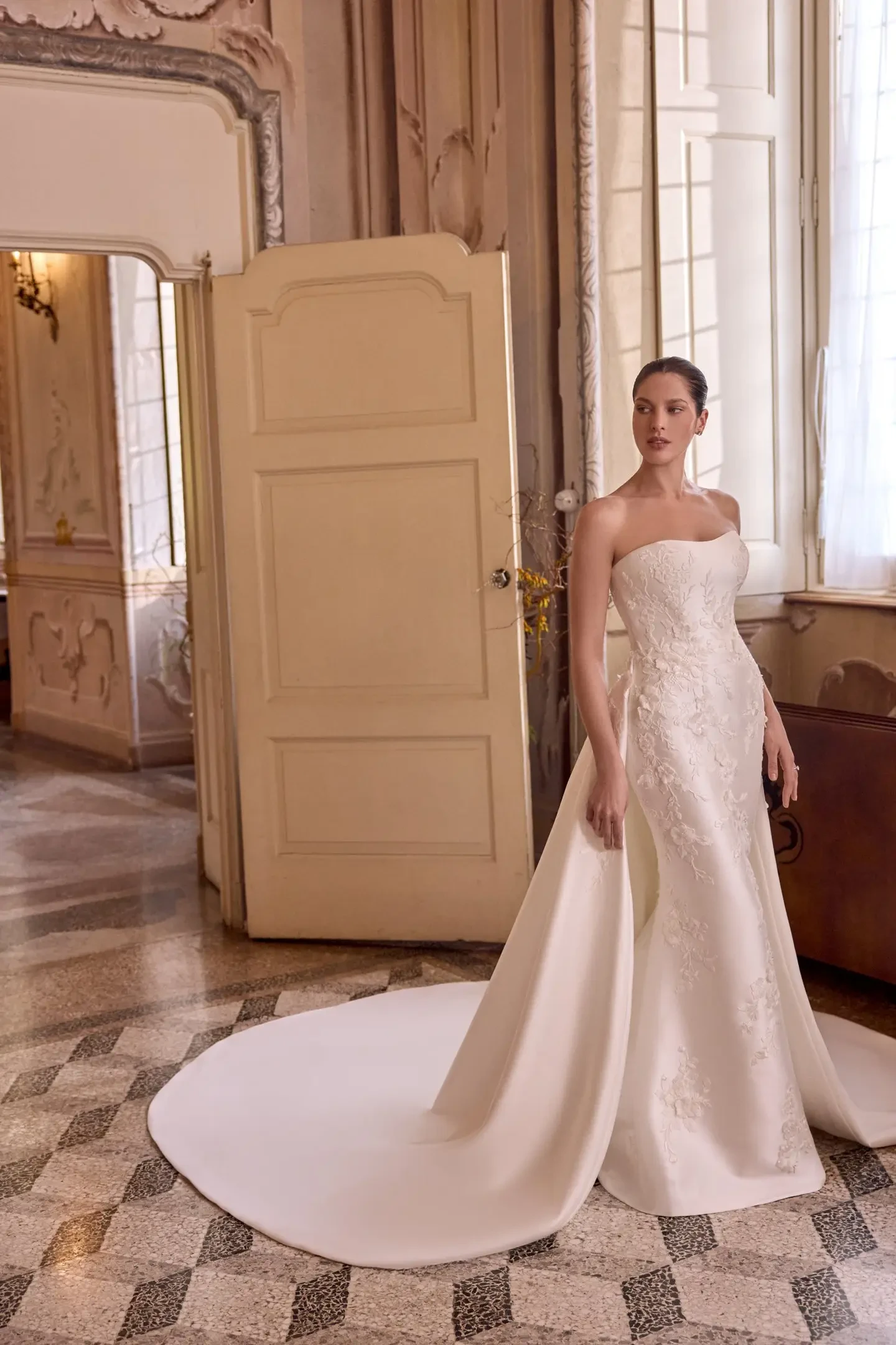 A woman in an elegant strapless white wedding gown with floral details stands in a grand, ornate room. The atmosphere is classic and sophisticated.